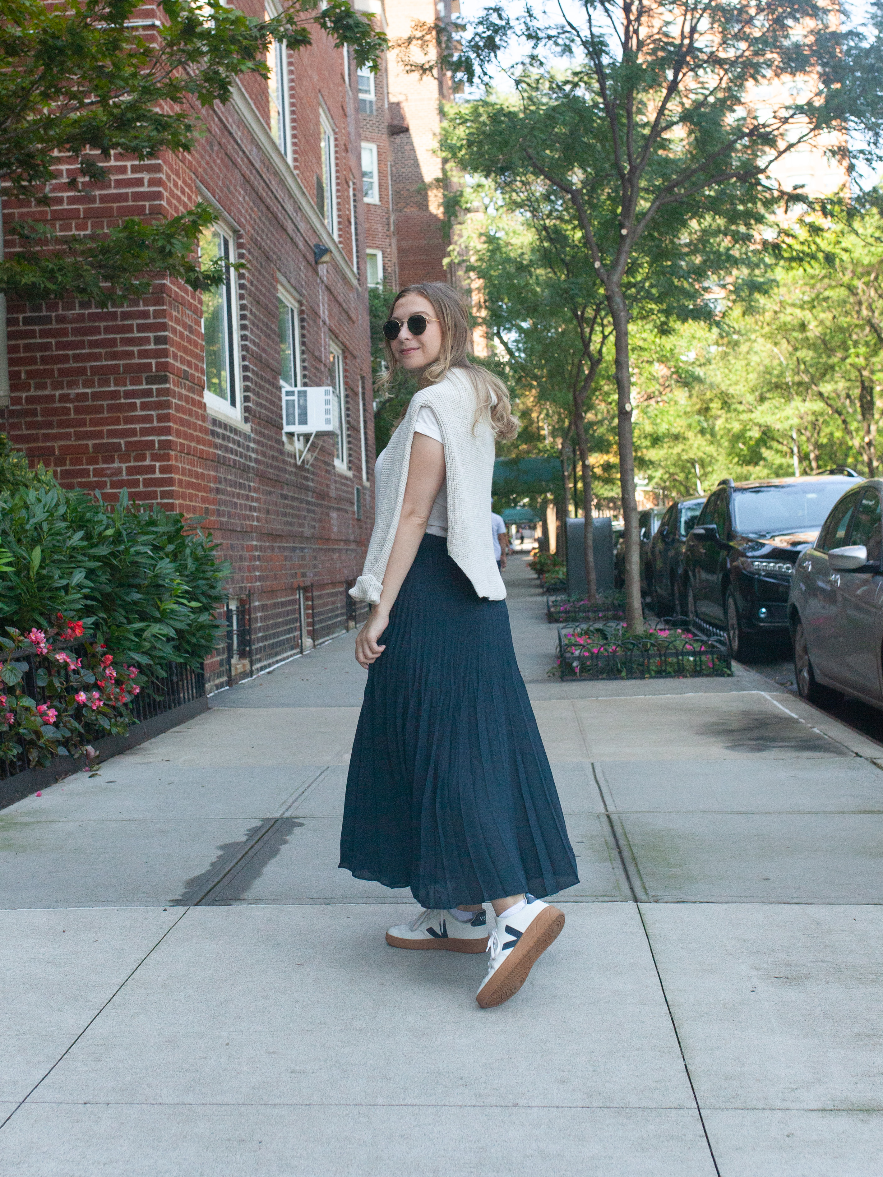 girl with blonde hair wearing navy blue maxi skirt, veja sneakers, white t-shirt and sunglasses with sweater draped over her shoulders