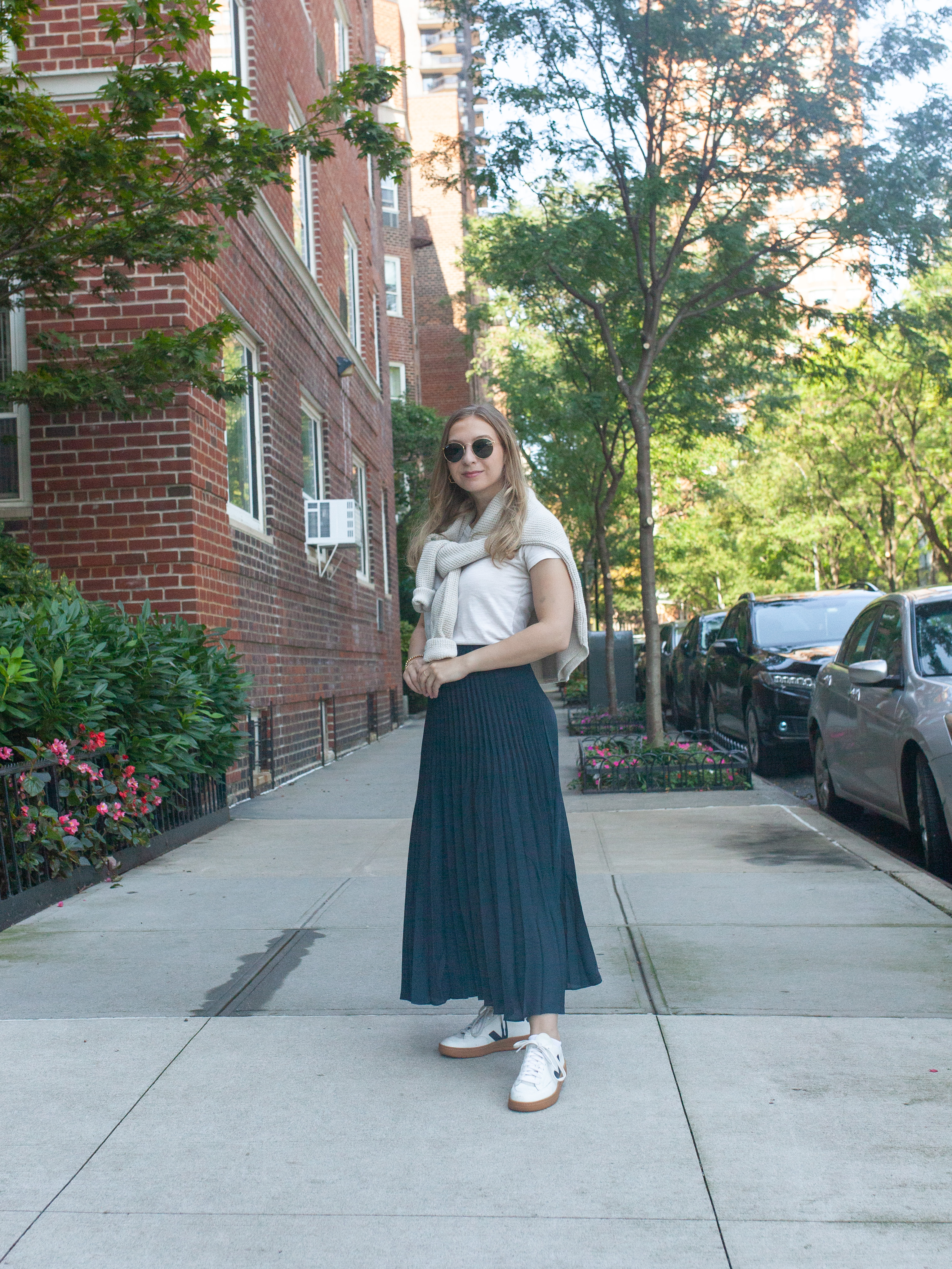 girl with blonde hair wearing navy blue maxi skirt, veja sneakers, white t-shirt and sunglasses with sweater draped over her shoulders