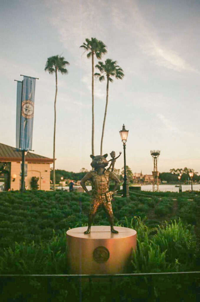 Bronze statue of Rocket from Guardians of the Galaxy with baby Groot on his shoulder in front of Palm Trees and the lake at Epcot in Walt Disney World