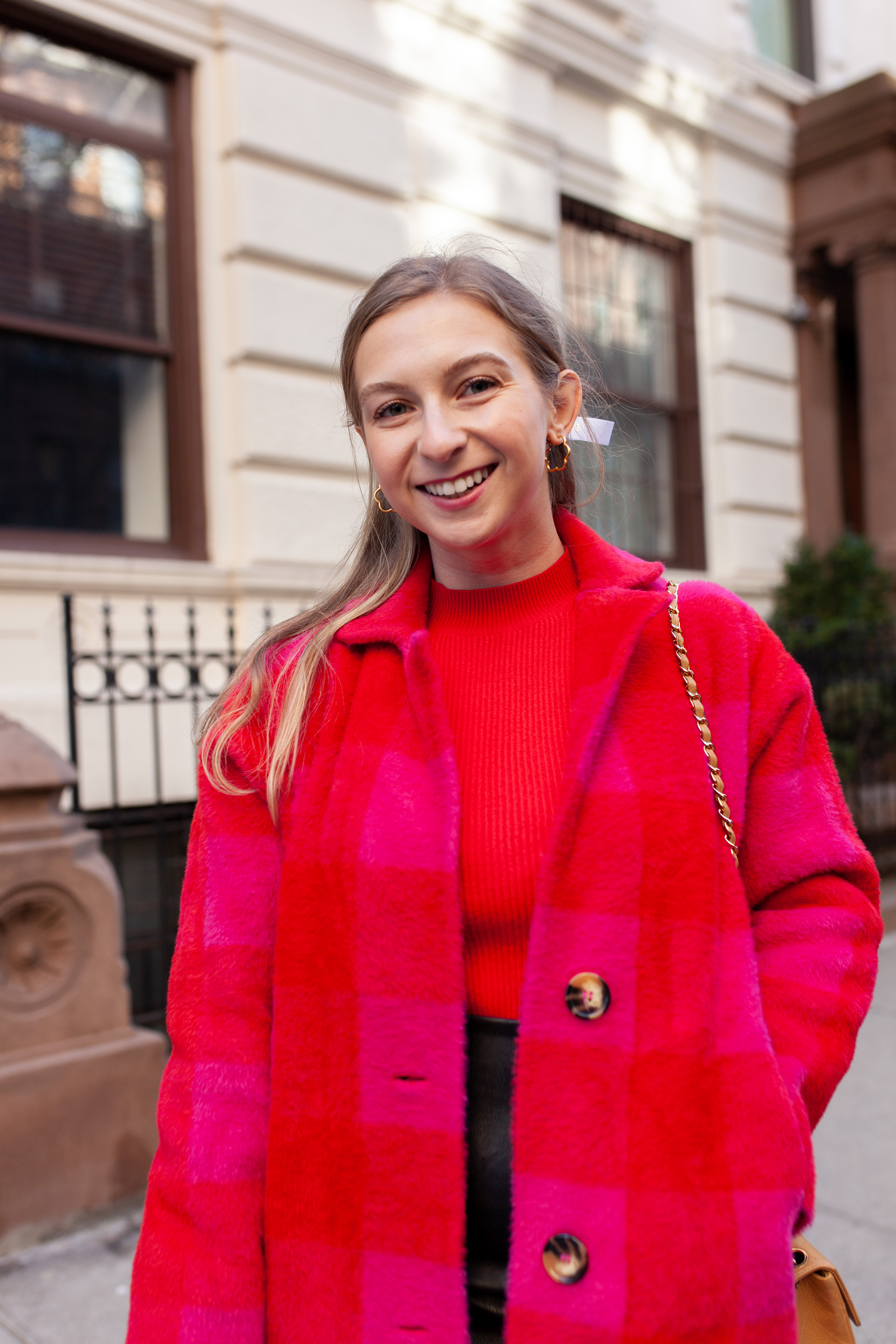 woman smiling at the camera, wearing pink and red check coat and red sweater, standing in front of nyc apartment building